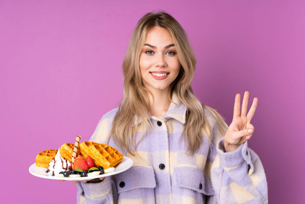 Неповнолітня російська дівчина з вафлями, ізольованими на пурпуровому фоні щасливою і порахованою трьома пальцями. - Фото, зображення