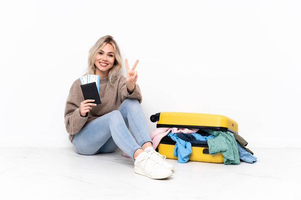 Подростковая блондинка с чемоданом, набитым одеждой, сидящая на полу, улыбаясь и показывая знак победы
 - Фото, изображение