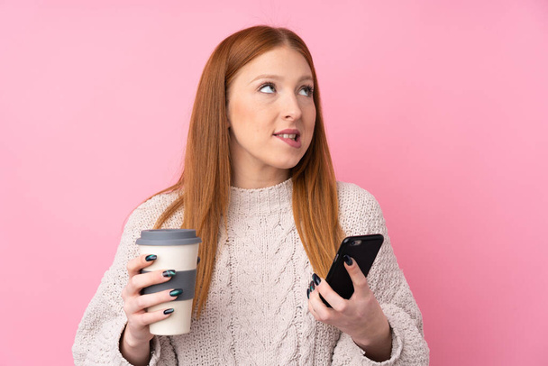 Jovem ruiva sobre fundo rosa isolado segurando café para tirar e um celular enquanto pensa em algo
 - Foto, Imagem