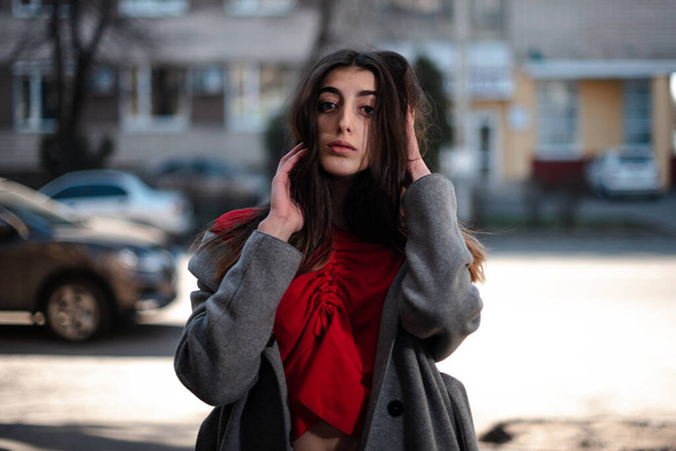 Girl in a red blouse and a gray cardigan on a blurry city background in early spring - Fotó, kép