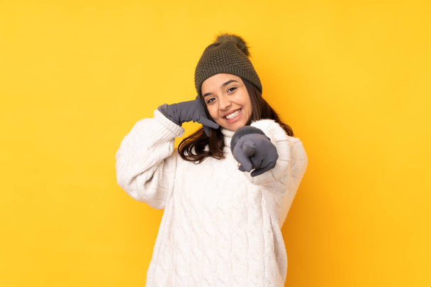Young woman with winter hat over isolated yellow background making phone gesture and pointing front - Φωτογραφία, εικόνα