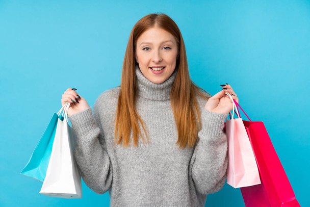 Молодая рыжая женщина на изолированном синем фоне держит сумки с покупками и улыбается - Фото, изображение