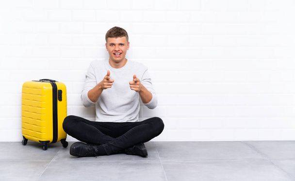 Young handsome man sitting on the floor with a suitcase points finger at you - Foto, afbeelding