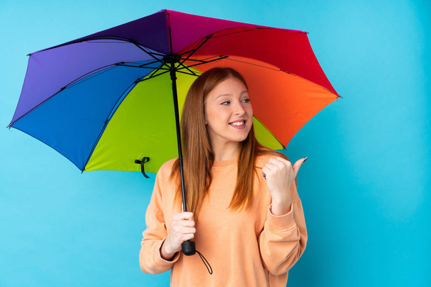 Junge rothaarige Frau hält einen Regenschirm über eine isolierte Wand und zeigt zur Seite, um ein Produkt zu präsentieren - Foto, Bild