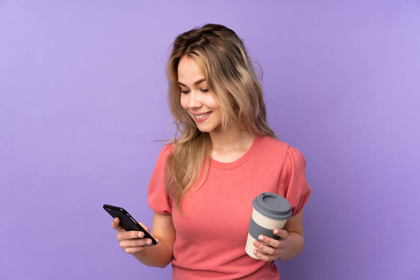 Teenager rusky dívka izolované na fialovém pozadí držení kávy odnést a mobilní - Fotografie, Obrázek