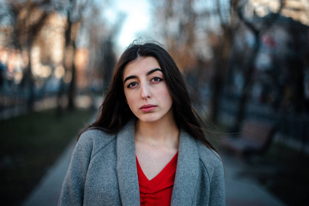 Girl in a red blouse and a gray cardigan on a blurry city background in early spring - Photo, image