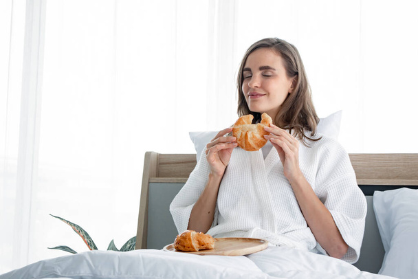 Красивая молодая кавказская женщина в пижаме, готовая к завтраку - круассаны по утрам после пробуждения на кровати в спальне дома. Молодая женщина кавказка ест хлеб после того, как я проснулся утром
. - Фото, изображение