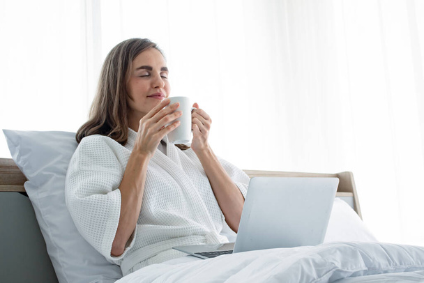 Jonge blanke vrouw in witte pyjama met wit met een kop koffie en drank en vind informatie van de laptop op een bed in een slaapkamer thuis. Vrouw die koffie drinkt en werkt op een laptop. - Foto, afbeelding
