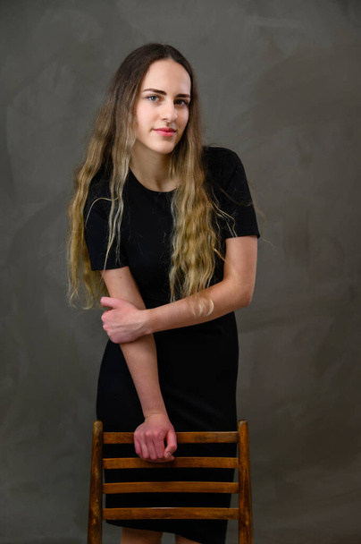 A model with long hair stands near a chair. Vertical portrait of a young pretty girl in a black dress on a gray background in the studio. - Фото, изображение