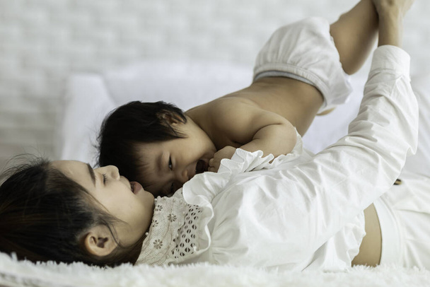 Happy Beauty mother Holding Cute Sweet Adorable Asian Baby wearing white dress smiling and playing with happiness emotional in cozy bedroom,Healthy Baby Concept,Selective Focus - Foto, Imagem