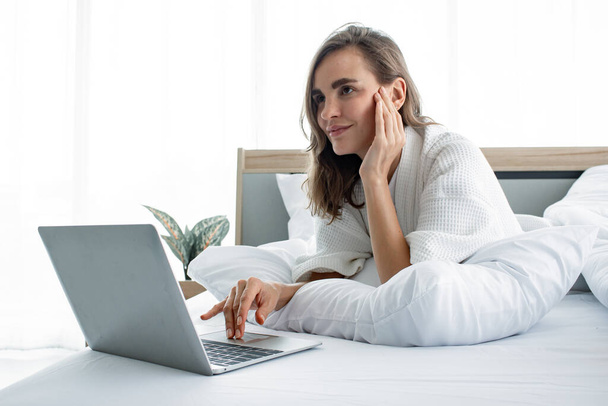 パジャマの美しい若い白人女性は、彼女の家で目を覚ました後、午前中に白いベッドの上でノートパソコンでソーシャルゲームをしています。女性はベッドのラップトップで誰かとチャット. - 写真・画像