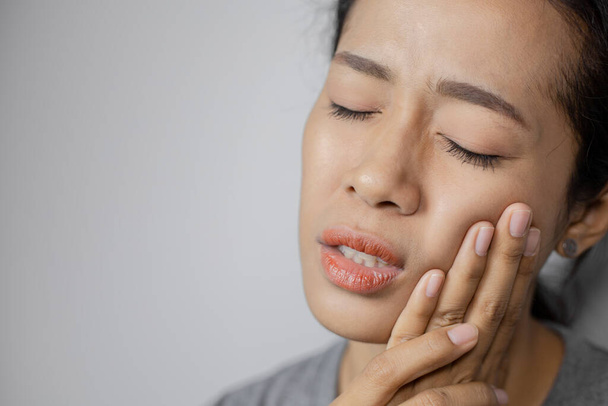 Woman put her hand on her cheek due to toothache. Asian  woman is suffering of toothache. Young woman with of toothache on a gray background. - Photo, Image