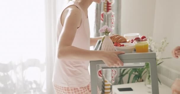 Beautiful woman carrying breakfast in bed for husband, kissing and sitting near him. Happy boyfriend waiting for his girlfriend holding tray with food. Concept of lifestyle and relationship. - Felvétel, videó