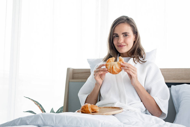 Kahvaltı yapmaya hazır beyaz pijamalı güzel bir kadın sabahları yataktaki yatak odasında ayçöreği ile uyanır. Ben sabah kalktıktan sonra beyaz bir kadın ekmek yiyor.. - Fotoğraf, Görsel