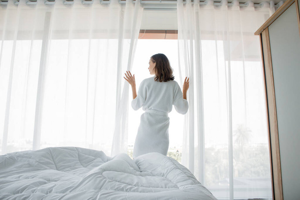 Νεαρή λευκή γυναίκα στέκεται ανοιχτή άσπρη κουρτίνα στο παράθυρο, το πρωί αφού ξύπνησε στην κρεβατοκάμαρα του σπιτιού της. Μια γυναίκα ξυπνάει με ένα φρέσκο και ανοίγει τις κουρτίνες στα παράθυρα.. - Φωτογραφία, εικόνα
