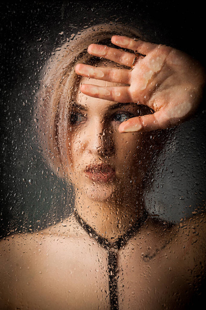 Portret of a blonde girl behind wet glass against a dark background. - Foto, afbeelding