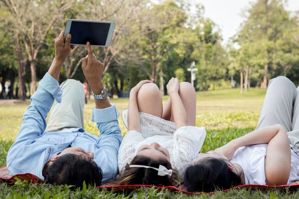 Asian family are using the tablet on the mat. Girls are learning from a tablet that parents are teaching. Parents and children are playing the tablet on the mat in the park. - Photo, Image