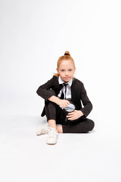 girl in a business black suit sits cross-legged and holds a magnifying glass - Фото, изображение
