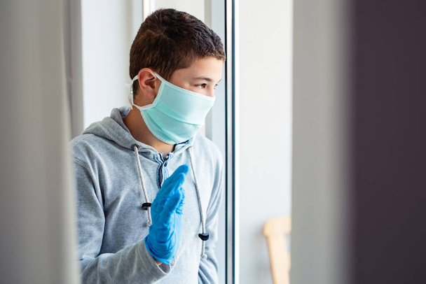 School age boy in a surgeon protection mask sitting by the window . Patient isolated to prevent infection.  - Zdjęcie, obraz