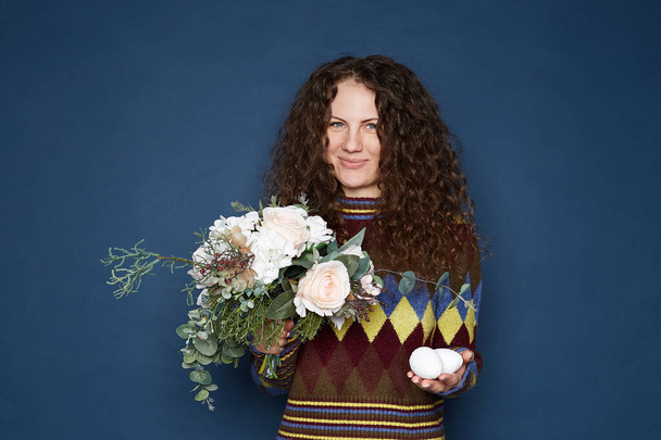 Wesołych Świąt Wielkanocnych. Urocza niebieskooka kręcone włosy kobieta gospodarstwa jaj i kwiatów, patrząc na aparat z czułym uśmiechem, pozowanie na ultramarynistycznej ścianie w pomieszczeniach, noszenie kolorowy przytulny sweter. - Zdjęcie, obraz
