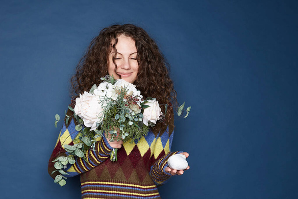 Happy spring family holiday concept. Authentic studio portrait of charming curly haired European female holding Easter eggs and flowers, smiling tenderly, isolated at home on blue wall background. - Photo, Image