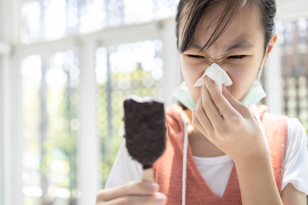 Doente menina asiática tem corrimento nasal e sopra o nariz em um papel tissue enquanto come muito sorvete, mulher com doença de resfriados, espirros, gripe, sintomas de infecção, problema respiratório
 - Foto, Imagem