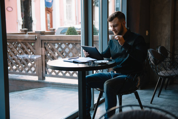 Knappe zakenman met behulp van een digitale tablet en het drinken van koffie tijdens het zitten in cafe in het centrum van de stad - Foto, afbeelding