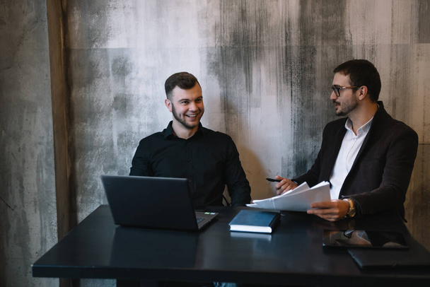 Two Businessmen In An Office Smiling At The Camera While Working Together Behind A Laptop Computer - Foto, afbeelding