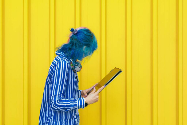 A young blue-haired female in a blue shirt holding a digital tablet device against a yellow wall - Foto, Imagem