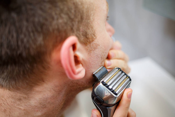 Muž si holí obličej elektrickou holicí stroječkou před zrcadlem. Dráždivost pro kůži. Koupací procedura - Fotografie, Obrázek