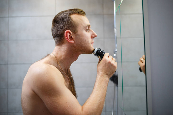 Ένας άντρας ξυρίζει το πρόσωπό του με ένα ηλεκτρικό ξυράφι μπροστά από έναν καθρέφτη. Ερεθισμός. Διαδικασία λουτρού - Φωτογραφία, εικόνα