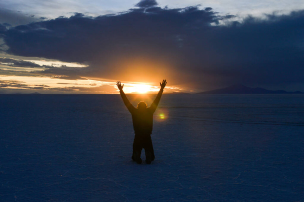 Homem ajoelhado de braços abertos e levantado no Salar de Uyuni, na Bolívia, ao pôr do sol
 - Foto, Imagem