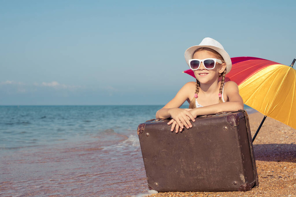 One happy little girl with suitcase sitting on the beach at the day time.  Kid having fun outdoors. Concept of summer vacation. - Foto, afbeelding