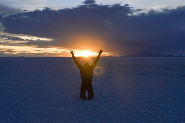 Mies polvistui kädet ylhäällä Salar de Uyunissa Boliviassa auringonlaskun aikaan
 - Valokuva, kuva
