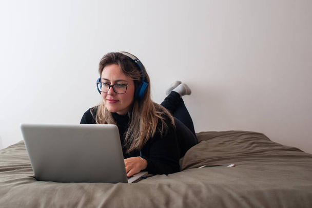 mulher loira deitada na cama usando o computador. ela está usando óculos e usando um fone de ouvido
 - Foto, Imagem