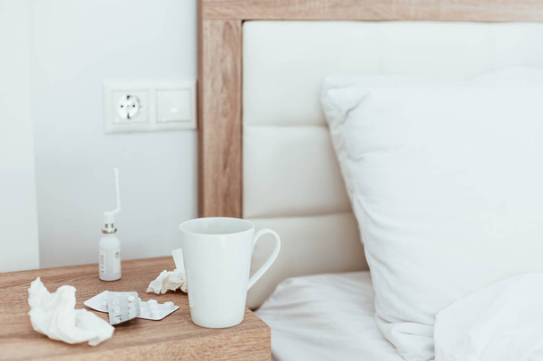 Medicines, pills, nose drops, tissues and hot drink cup on bedside table in modern apartments. Being sick, unwell, flu, illness symptoms. Coronavirus home treatment concept. Lifestyle image copy space - 写真・画像