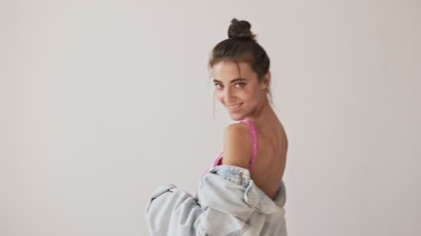 Playful stylish pretty brunette woman in swimsuit and jacket dancing and looking at the camera over grey background - Záběry, video