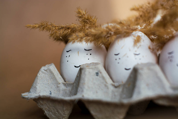 Happy Easter and white eggs in corton tray with cute faces in wreaths made of spikelets. - Photo, Image