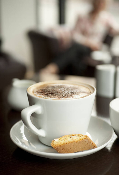A Cappuccino with chocolate dusting on the foam with an italian Biscotti perched on the rim of the saucer in a cafe style setting - Photo, Image
