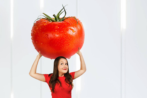 Young hunger pretty funny woman holds huge and large red tomato with drops water. Girl walks with big heavy vegetable. - Photo, Image