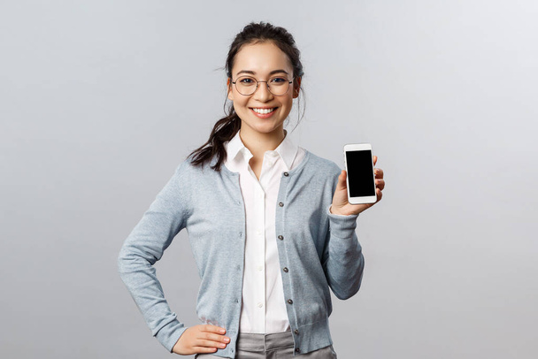 Technológia, online és mobil életmód koncepció. Vidám ázsiai nő szemüvegben be új alkalmazás, internetes áruház link, mutatja okostelefon kijelző elégedett mosollyal - Fotó, kép