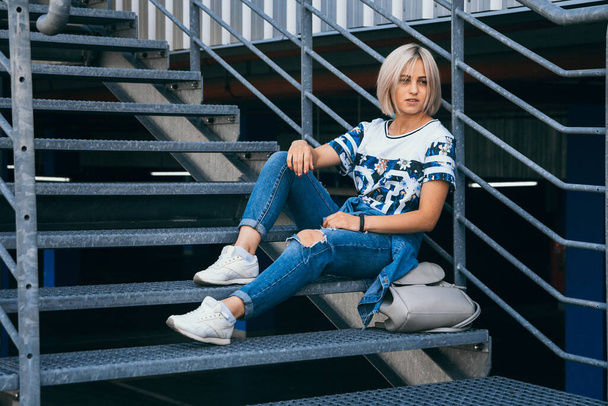 Beautiful girl with short white hair dressed in jeans in urban style sits on the metal stairs. Place for text - Photo, Image