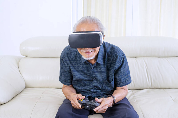 Retrato del hombre mayor sentado en el sofá mientras juega a videojuegos con gafas de realidad virtual y joystick
 - Foto, imagen