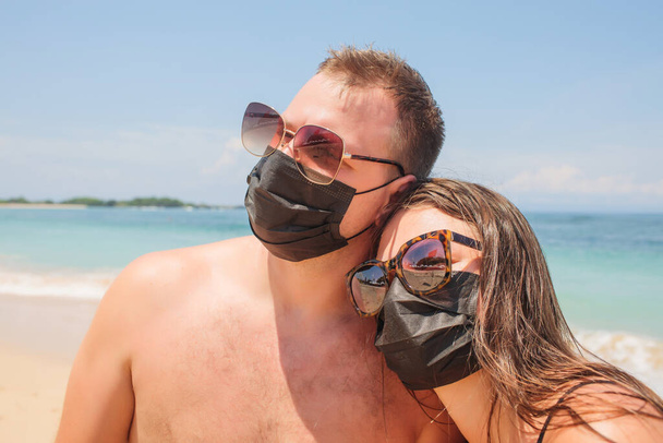 Joven pareja interracial usando máscara respiratoria de pie junto a la playa protegiéndose de la infección viral, virus wuhan, virus corona durante las vacaciones
. - Foto, Imagen