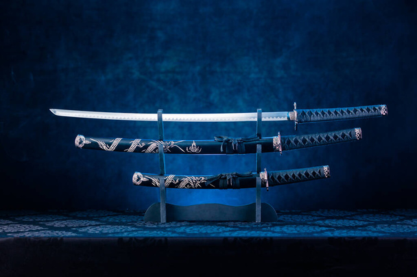Drie zwaarden op standaard, katanaklemmet blootgesteld, blauw licht achter - Foto, afbeelding