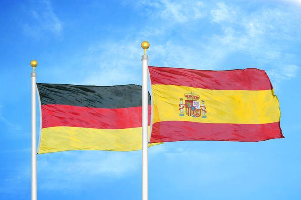 Németország és Spanyolország két zászló zászlórúd és kék felhős ég háttér - Fotó, kép