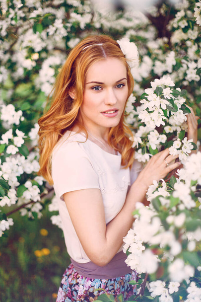 Model with red hair posing in a blooming apple tree. - Zdjęcie, obraz