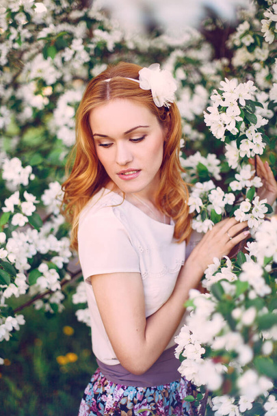 Model with red hair posing in a blooming apple tree. - Φωτογραφία, εικόνα