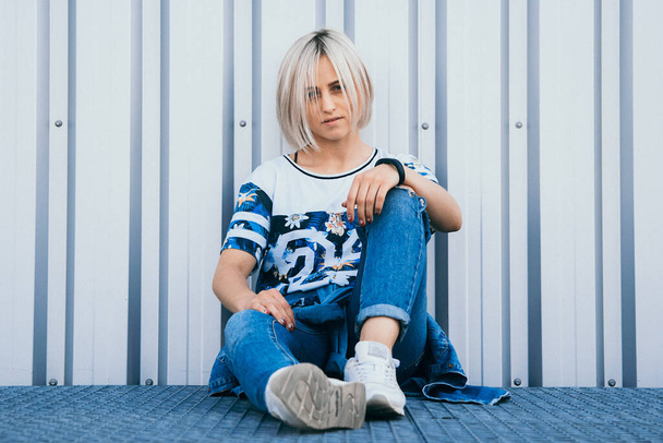 Beautiful girl with short white hair dressed in jeans in urban style sits on the background of a white wall. A look at the camera - Foto, immagini