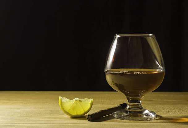 A glass glass of cognac on a wooden table, a slice of lemon and a cigar Free space for text on the side. - Foto, immagini